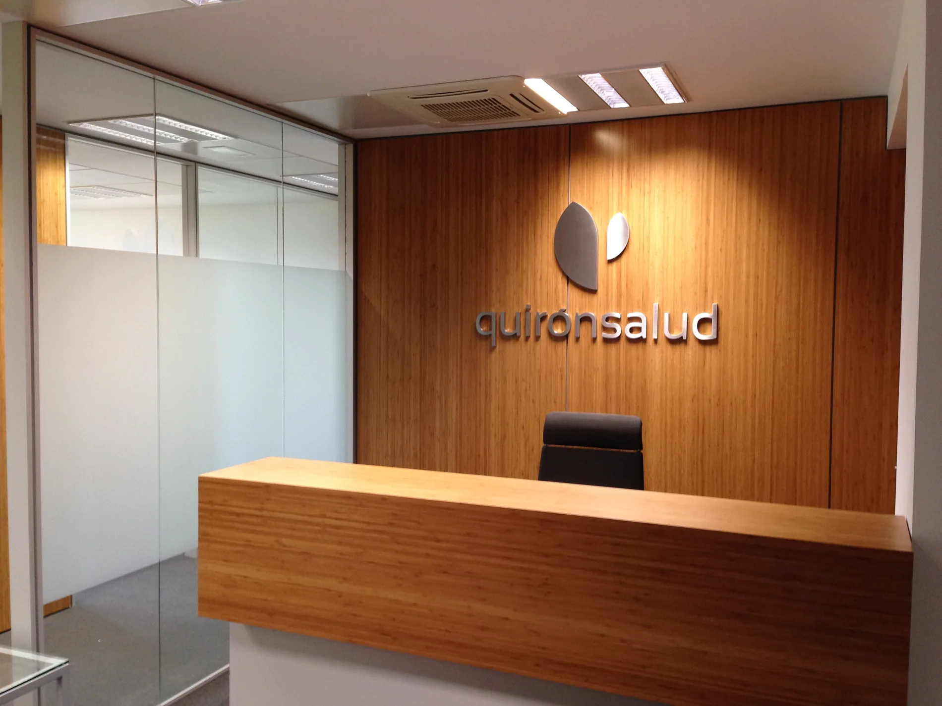 Recepción de oficinas de Quirónsalud con el logotipo en letras corpóreas de metal sobre el frente de madera y la mampara de vidrio con vinilo efecto ácido